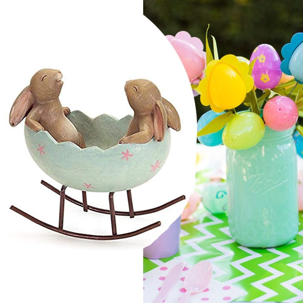 Easter figurine of two bunnies in a cracked egg cradle on a rocking base, with Easter eggs in a jar in the background.