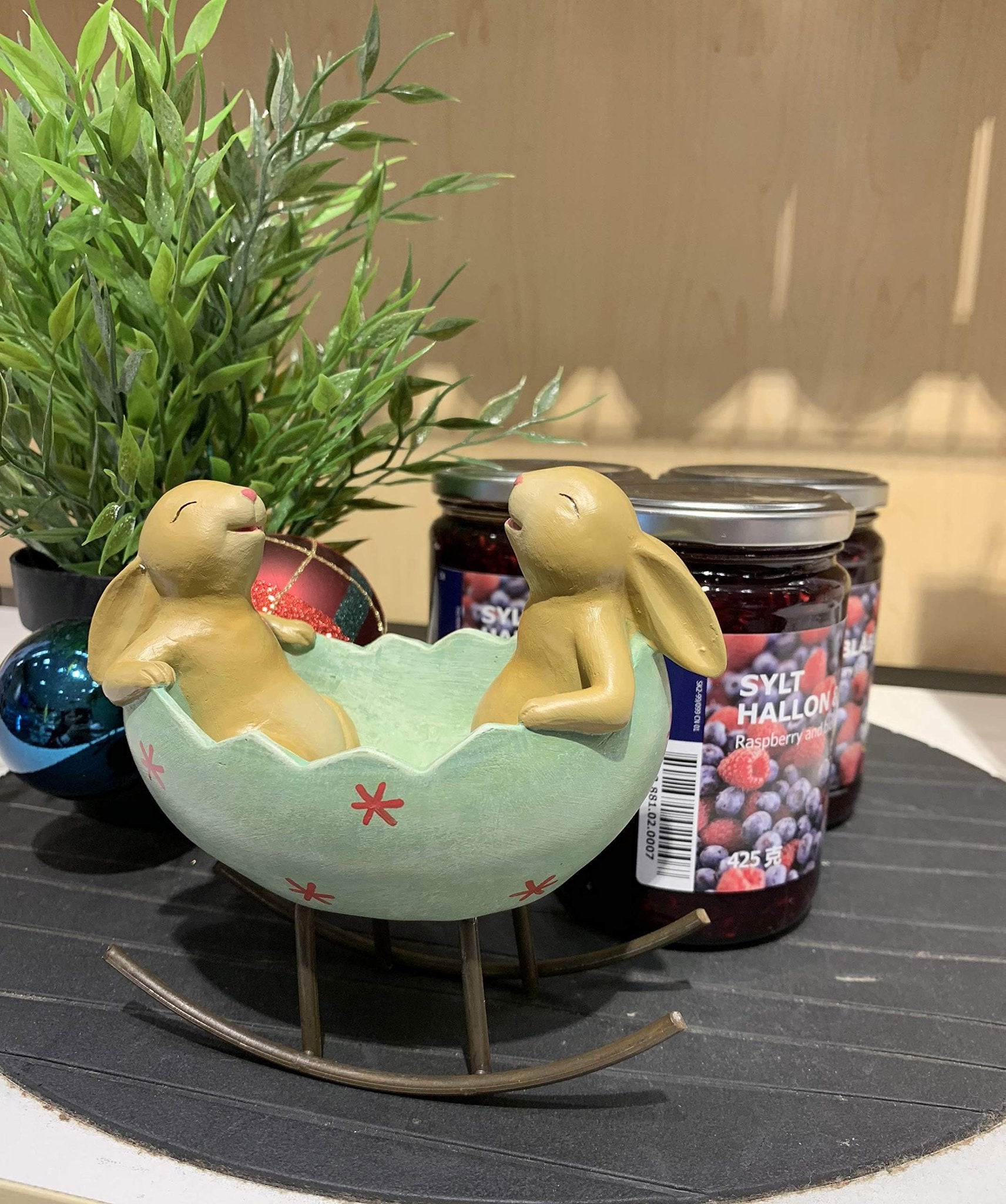 Easter figurine of two bunnies in a green egg cradle with red asterisks, next to a potted plant and jars of jam.