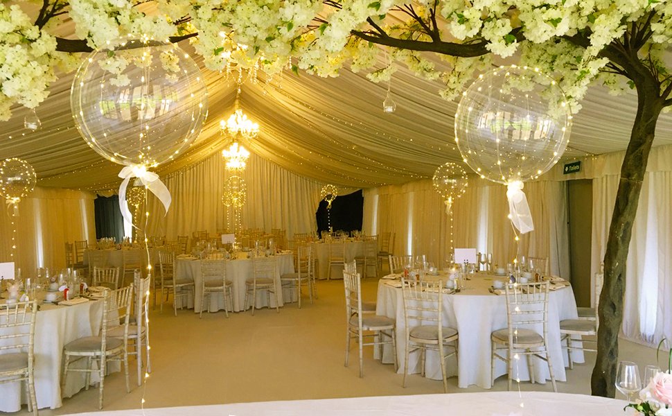 Clear LED light-up bobo balloons with warm white string lights decorating a wedding reception tent with tables and chairs.