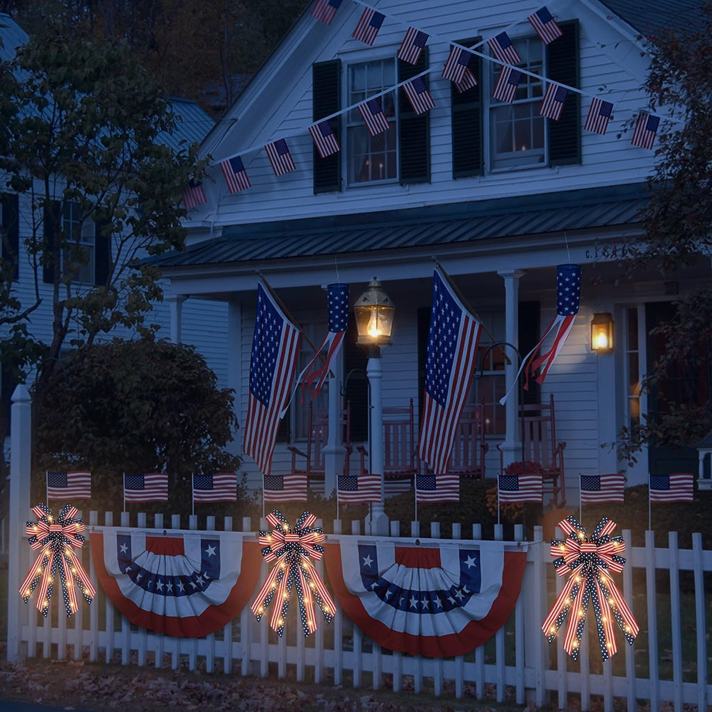 Patriotic burlap bows with warm white lights decorating a white picket fence in front of a house at dusk.