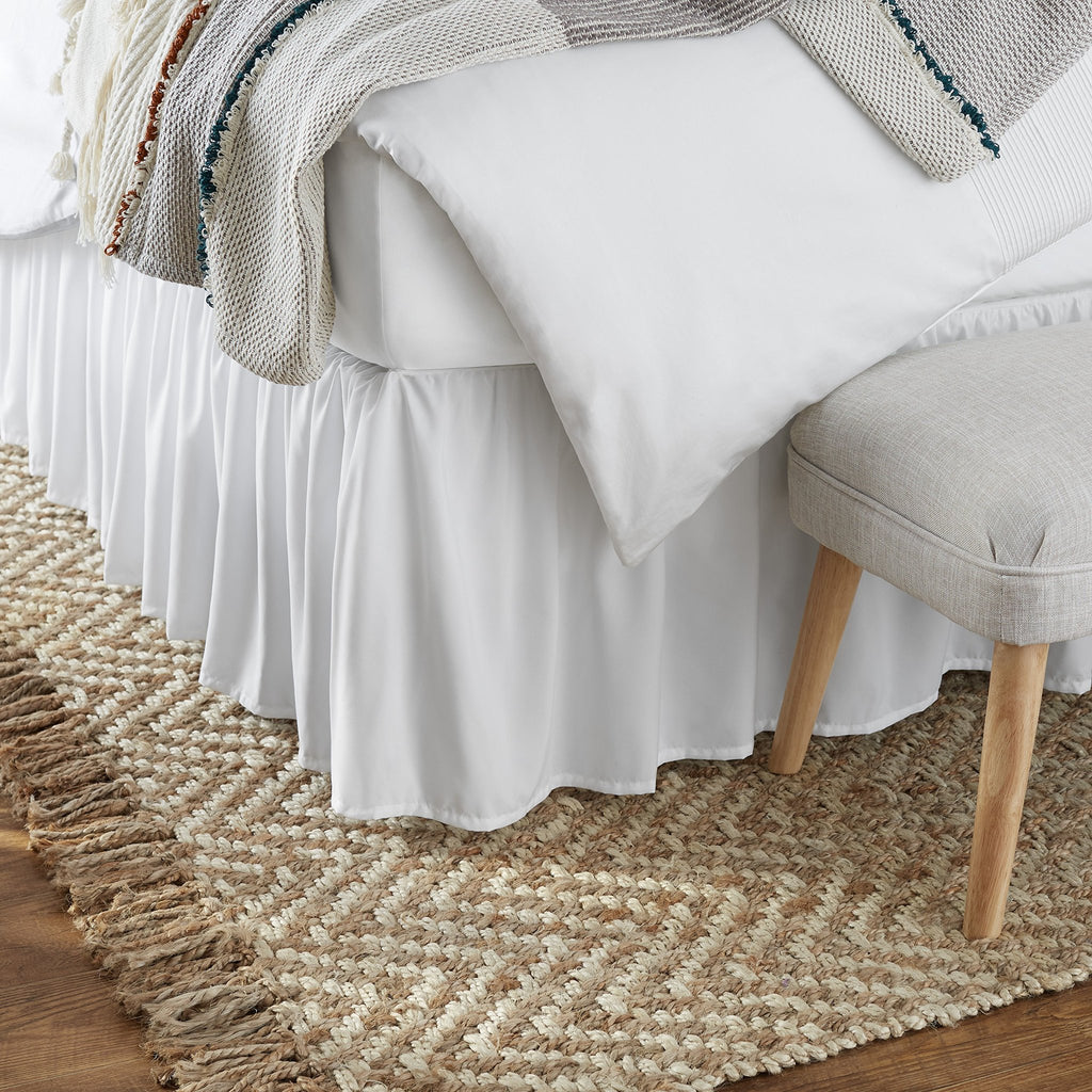 Close-up of a white ruffled bed skirt with a textured throw blanket draped over the bed and a small upholstered bench at the foot of the bed.