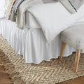 Close-up of a white ruffled bed skirt with a textured throw blanket draped over the bed and a small upholstered bench at the foot of the bed.