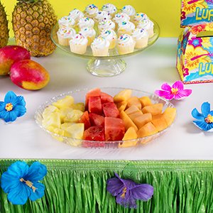 Tropical party table with fruit platter, cupcakes, and a green grass table skirt decorated with hibiscus flowers.