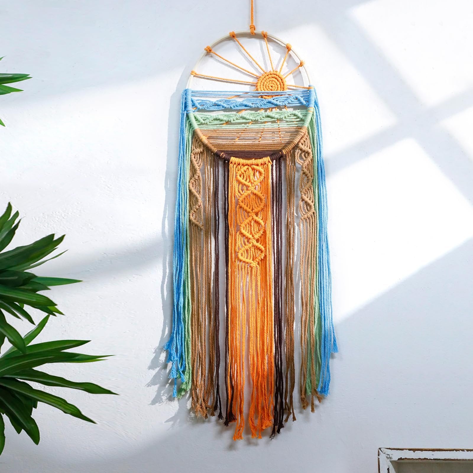 Macrame sun and wave wall hanging with colorful tassels, displayed on a white wall with plant and shadow accents.