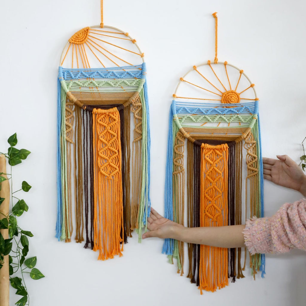 Two macrame sun dream catchers with colorful yarn strands hanging on a white wall, one held by hands.