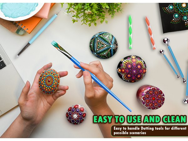 Hands holding a painted mandala stone while a brush with green paint is poised to add more dots. Mandala painting tools and decorated stones are scattered around.