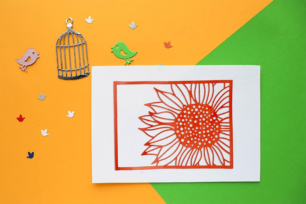 Orange sunflower cutting die on white cardstock with decorative bird and birdcage elements on orange and green background