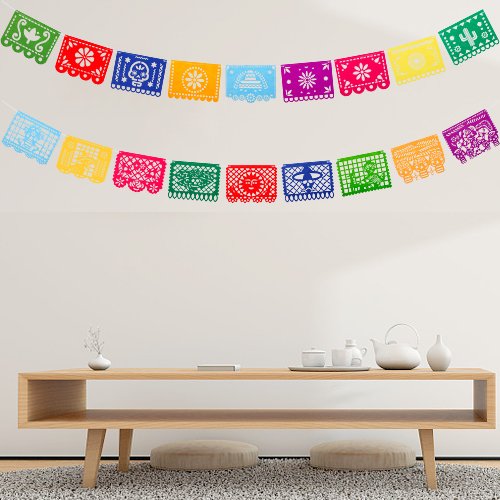 Mexican fiesta papel picado banners with 18 colorful felt flag designs hanging on a white wall above a wooden table.