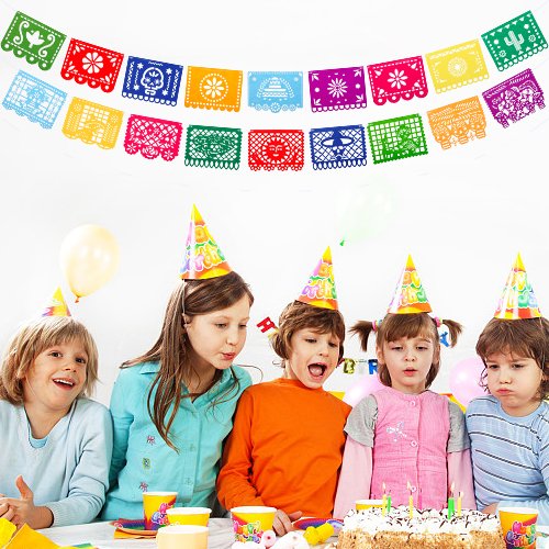 Children wearing party hats gathered around a birthday cake with colorful Papel Picado banners overhead.
