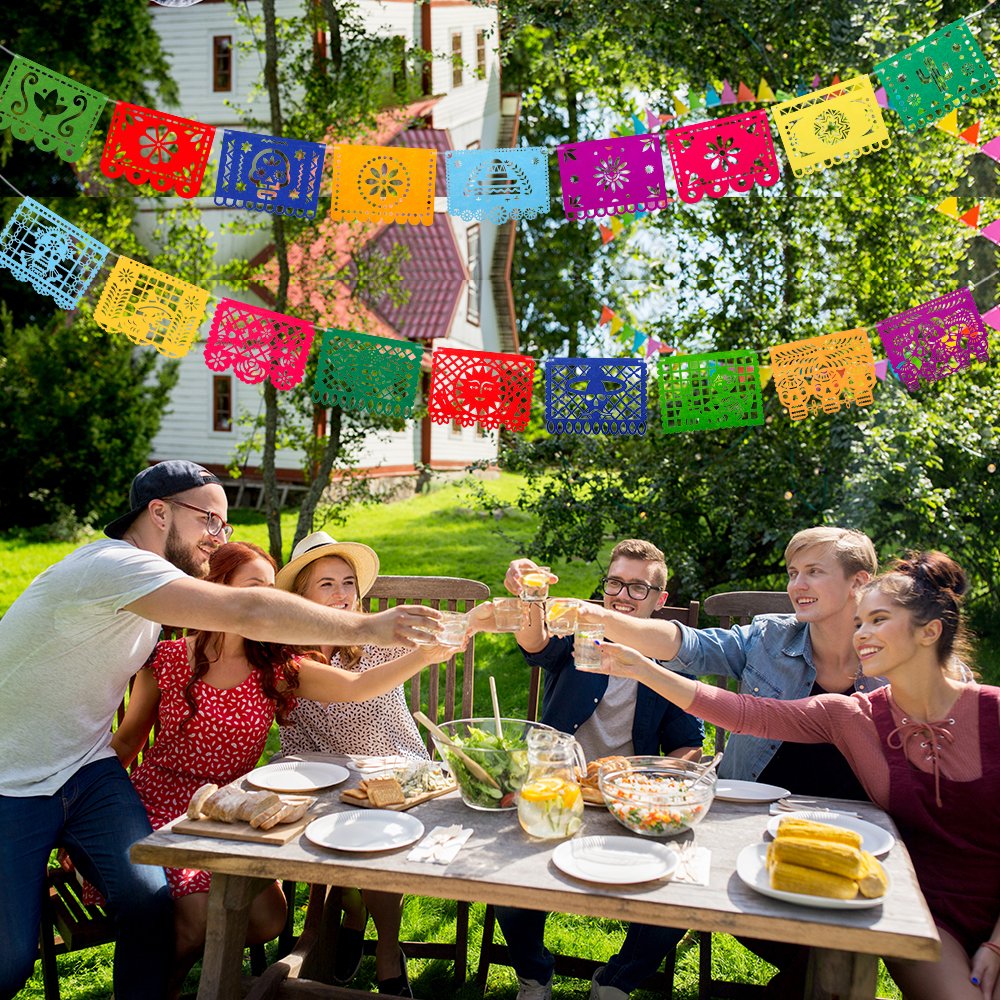 Group of friends toasting drinks at a picnic table with colorful Mexican fiesta papel picado banners overhead