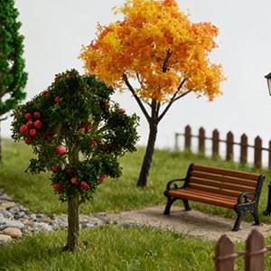 Miniature apple tree with red fruit and a miniature autumn tree in a diorama setting with a bench and fence.