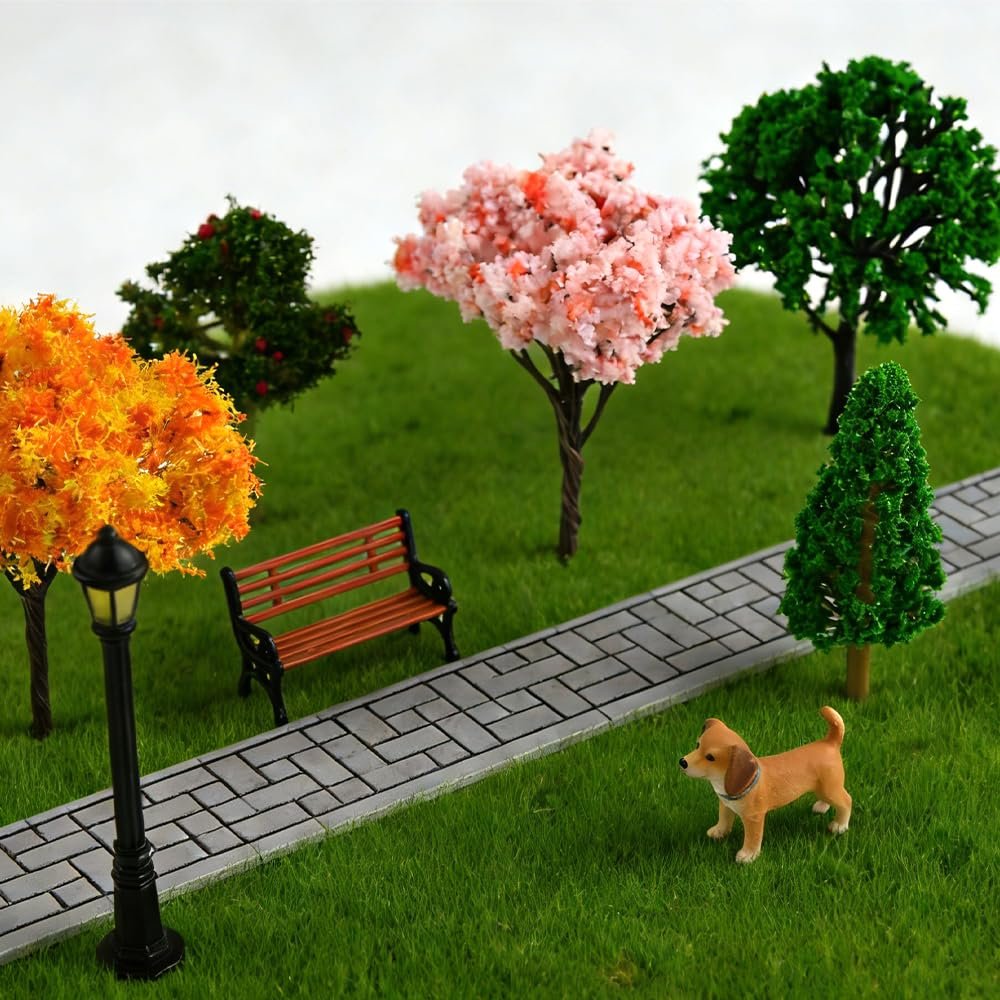 Miniature park scene with assorted artificial trees, a bench, lamppost, and a small dog on a paved path.
