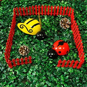 Miniature red picket fence enclosing a fairy garden scene with a bee, ladybug, and pinecones on artificial grass.