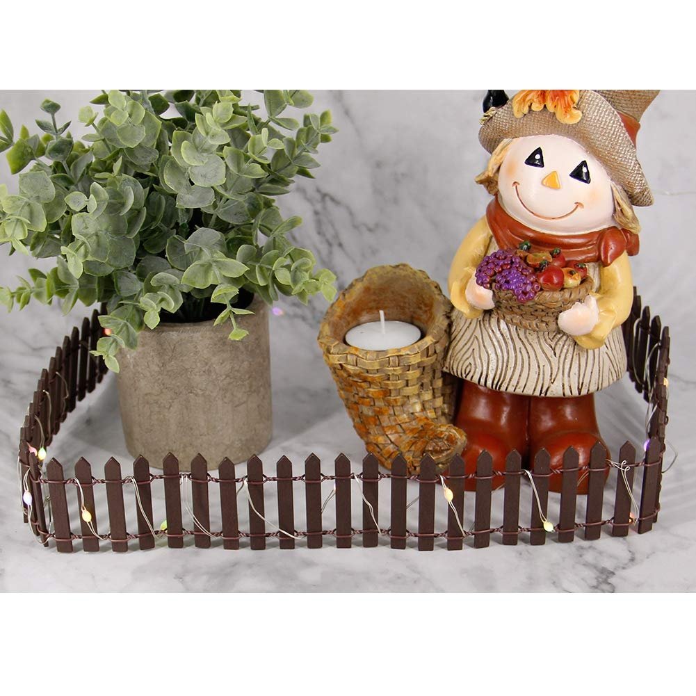 Miniature brown picket fence with fairy lights surrounding a scarecrow figure, candle holder, and potted plant.