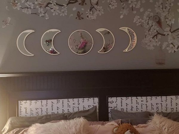 Five acrylic moon phase mirrors arranged on a wall above a bed, with floral wall decals in the background.