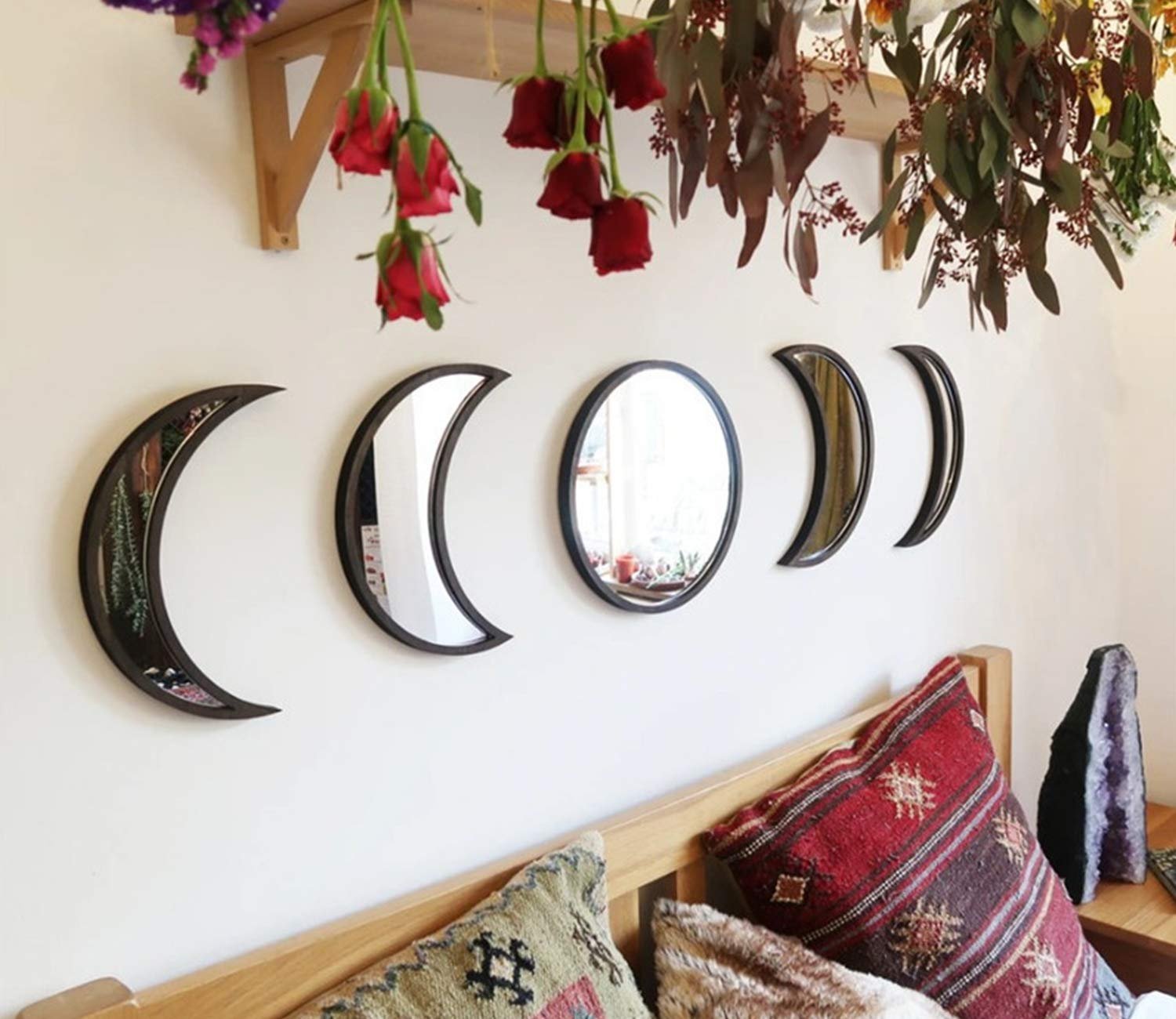 Five moon phase mirrors arranged on a wall above a bed with decorative pillows and hanging flowers.