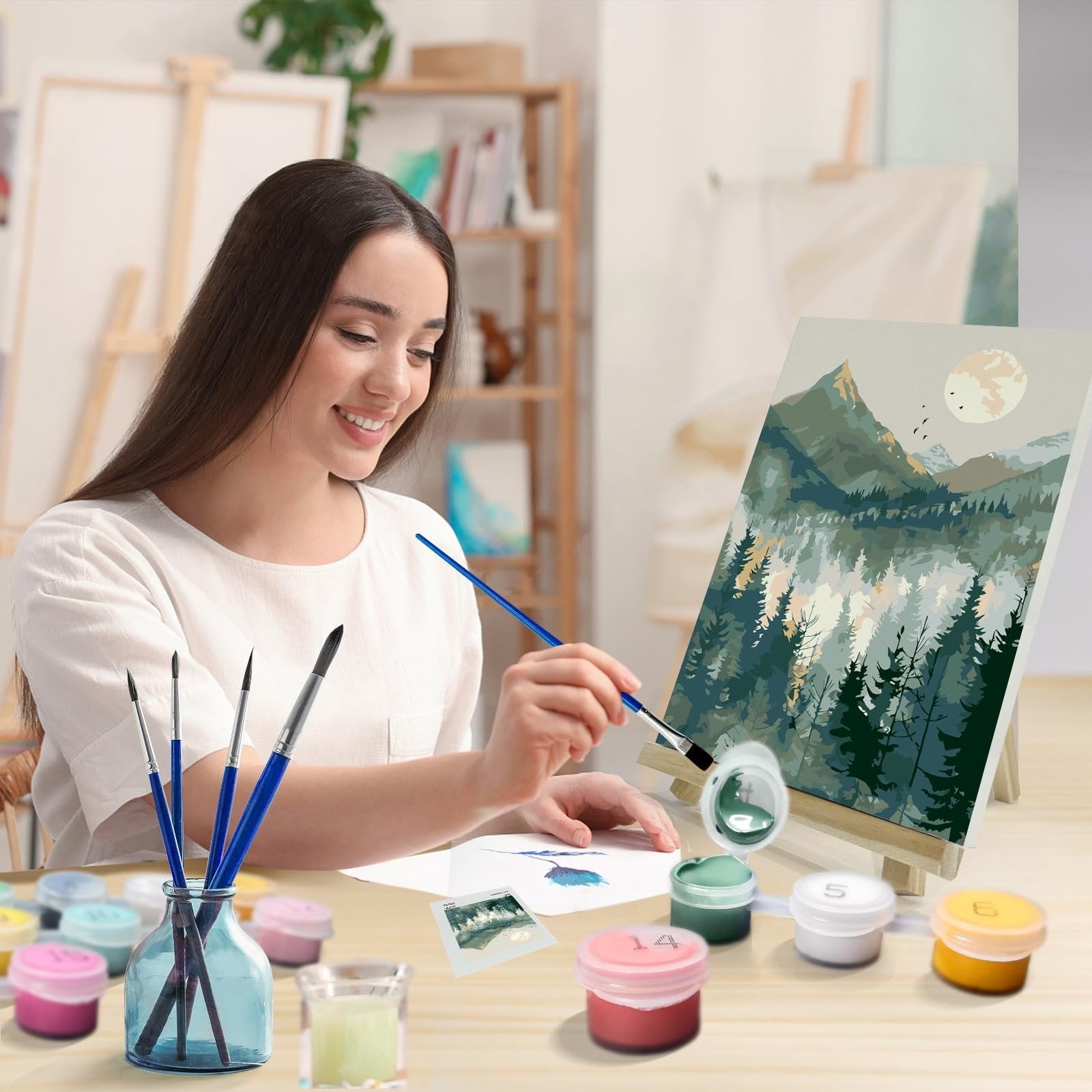 Woman painting a mountain landscape by number kit with frame, easel, brushes and acrylic paints on a table