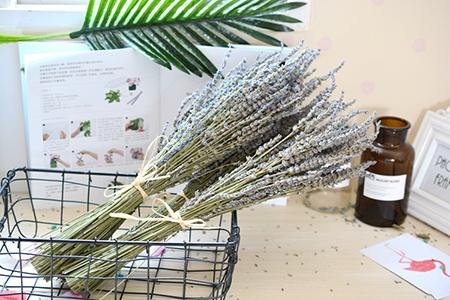 Two bundles of dried lavender flowers tied with twine, resting in a black wire basket next to an open book and a brown glass bottle.