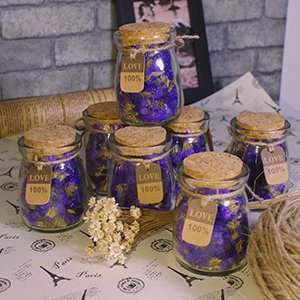 Seven small glass jars filled with purple potpourri, each with a cork lid and a small tag reading 'LOVE 100%', arranged on a surface with Paris-themed print.