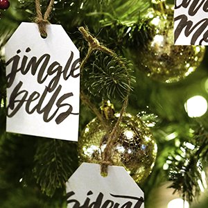 Christmas tree ornaments with handwritten text tags and gold baubles hanging on branches with lights.