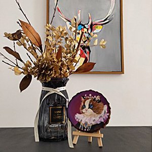 Painted wooden slice of a cat in a crown, displayed on a mini easel next to a vase of dried flowers.
