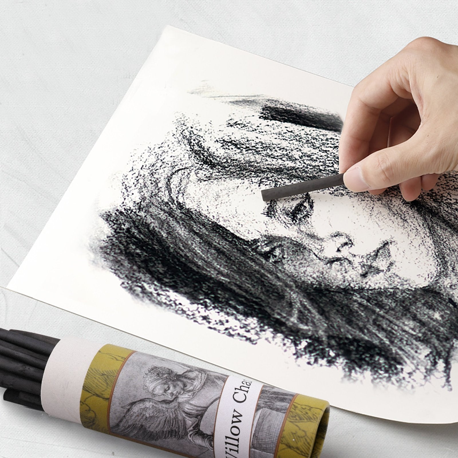 Artist's hand sketching a portrait with willow charcoal sticks on paper, with a tube of charcoal sticks in the foreground.