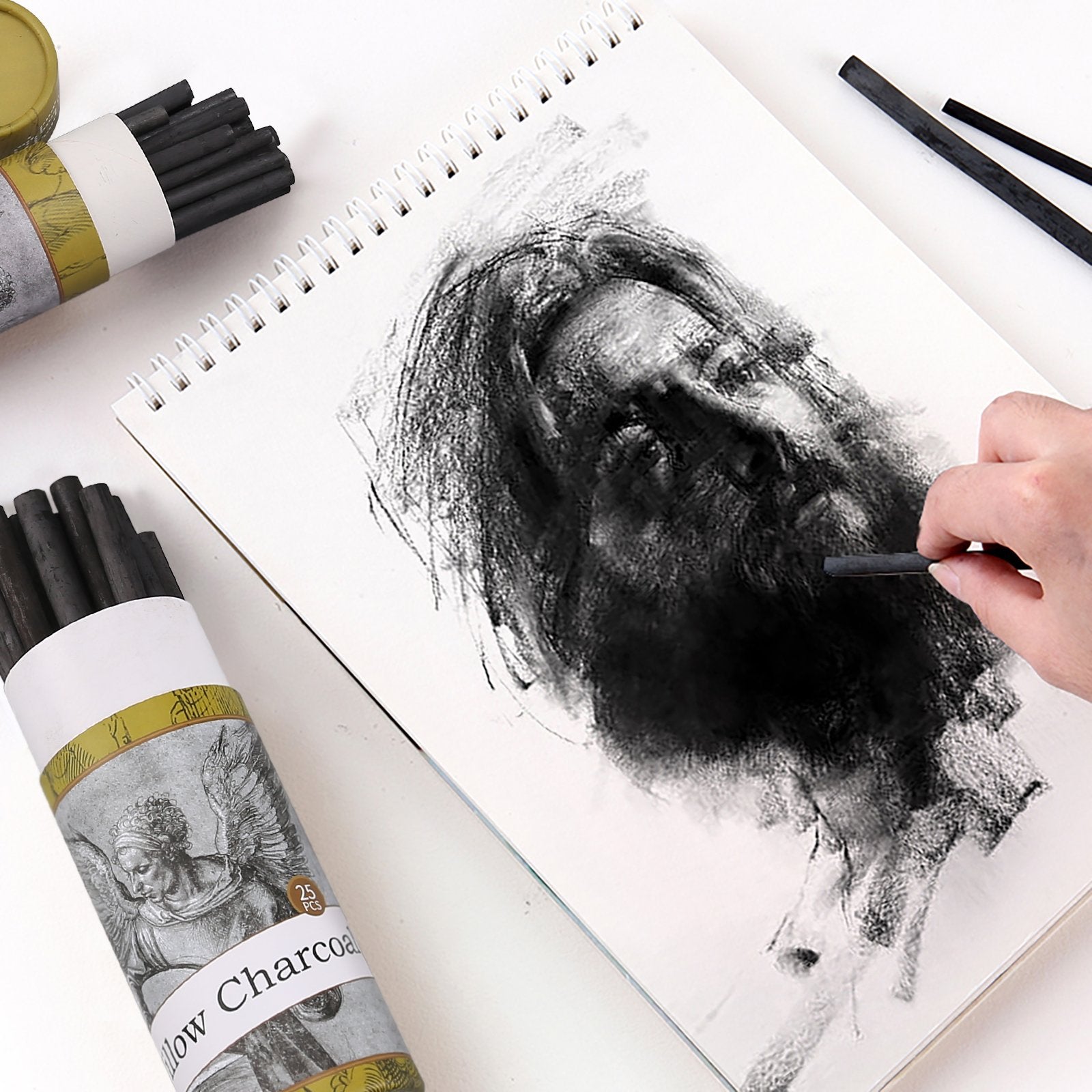Artist sketching a portrait with willow charcoal sticks on a spiral-bound sketchbook page, with charcoal tubes in foreground.