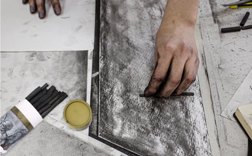 Artist's hands drawing with charcoal sticks on paper, with a pack of charcoal sticks and a small notebook nearby.