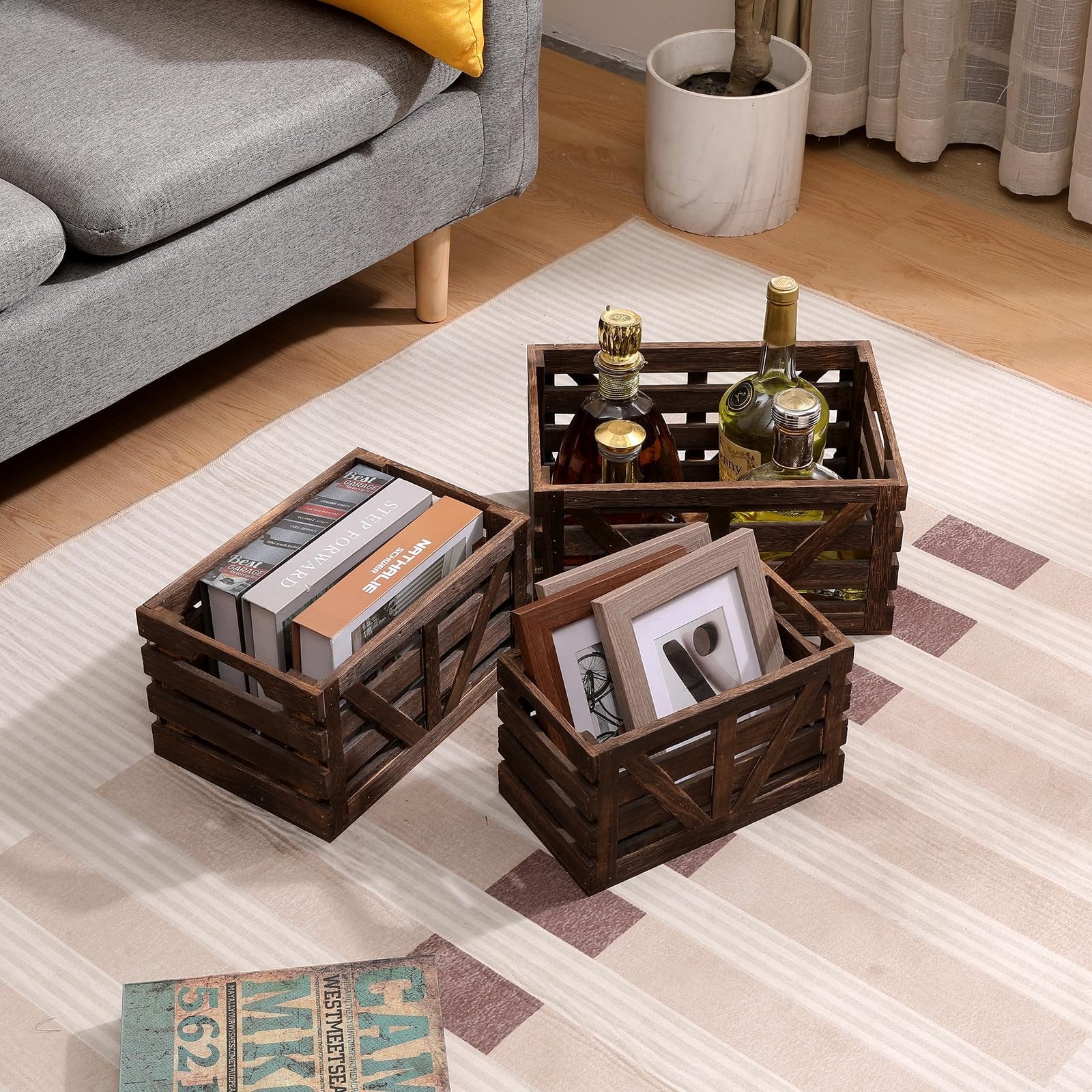 Three wooden storage crates arranged on a rug, one filled with books, another with picture frames, and the third with bottles.