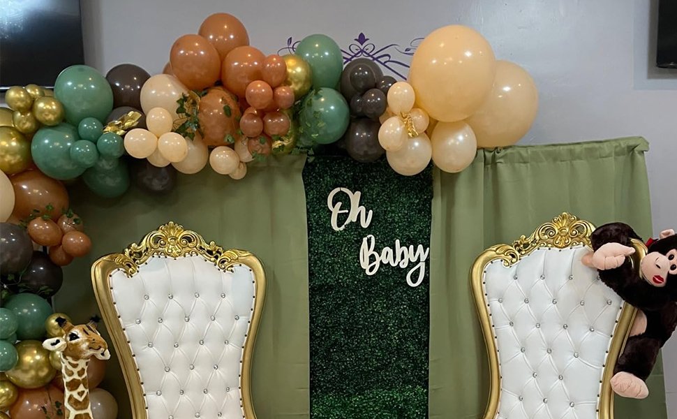 Jungle-themed baby shower decor with balloon arch, two ornate chairs, and 'Oh Baby' sign on a green hedge wall.