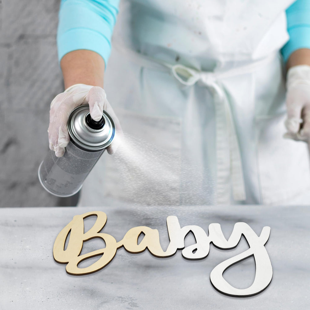 Person in gloves spraying a can onto a wooden 'Baby' sign on a gray surface.