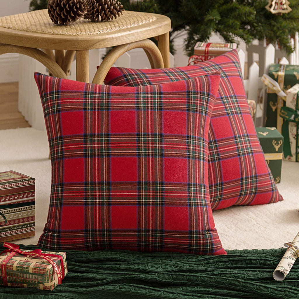 Two red and green plaid Christmas throw pillow covers on a cream rug with pine cones and gifts.