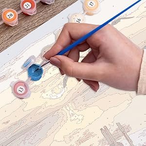 Hand holding a blue paintbrush applying blue paint to a paint by numbers canvas with numbered paint pots nearby