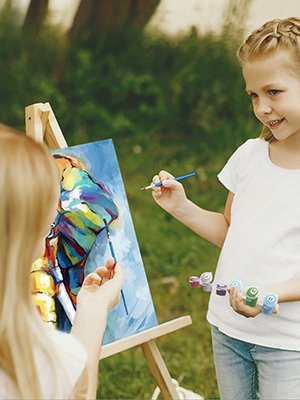 Two girls painting a colorful abstract design on canvas outdoors with brushes and paint pots