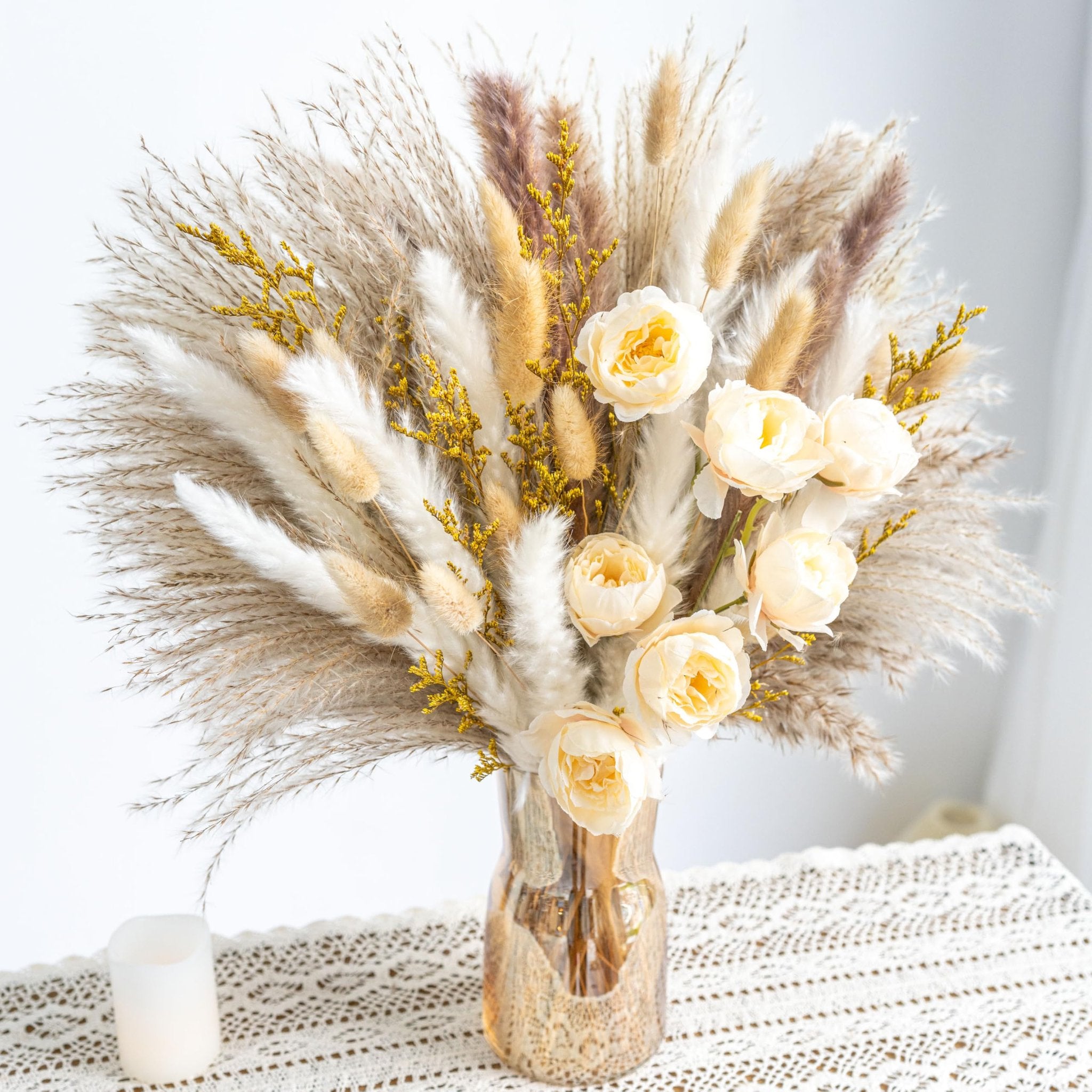 Boho dried flower and pampas grass bouquet in a textured amber vase on a lace tablecloth with a candle.