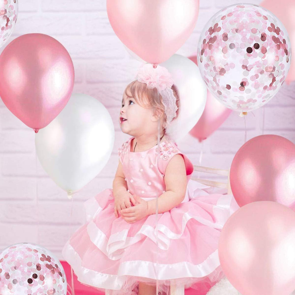 Baby girl in pink dress surrounded by pink, white, and confetti balloons against a white brick background.
