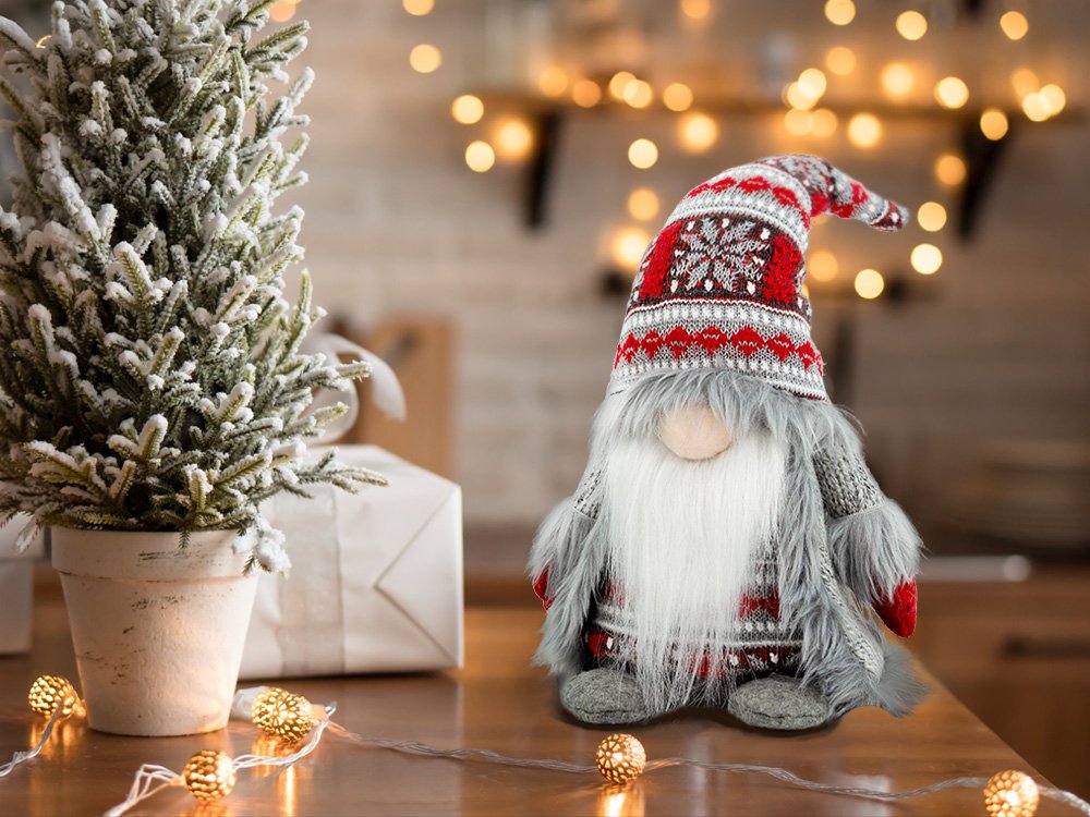 Plush Christmas gnome decoration with gray knit outfit and festive hat, next to a small frosted tree and gift.