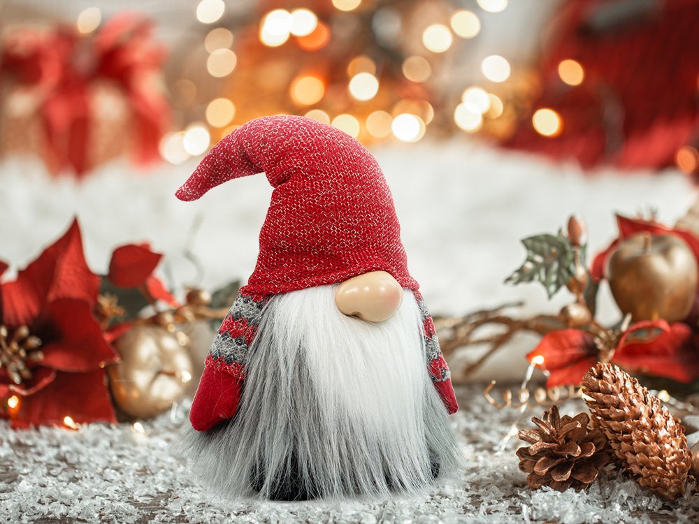 Plush Christmas gnome with a red knit hat and gray beard, set against a festive, blurred background with lights and decorations.