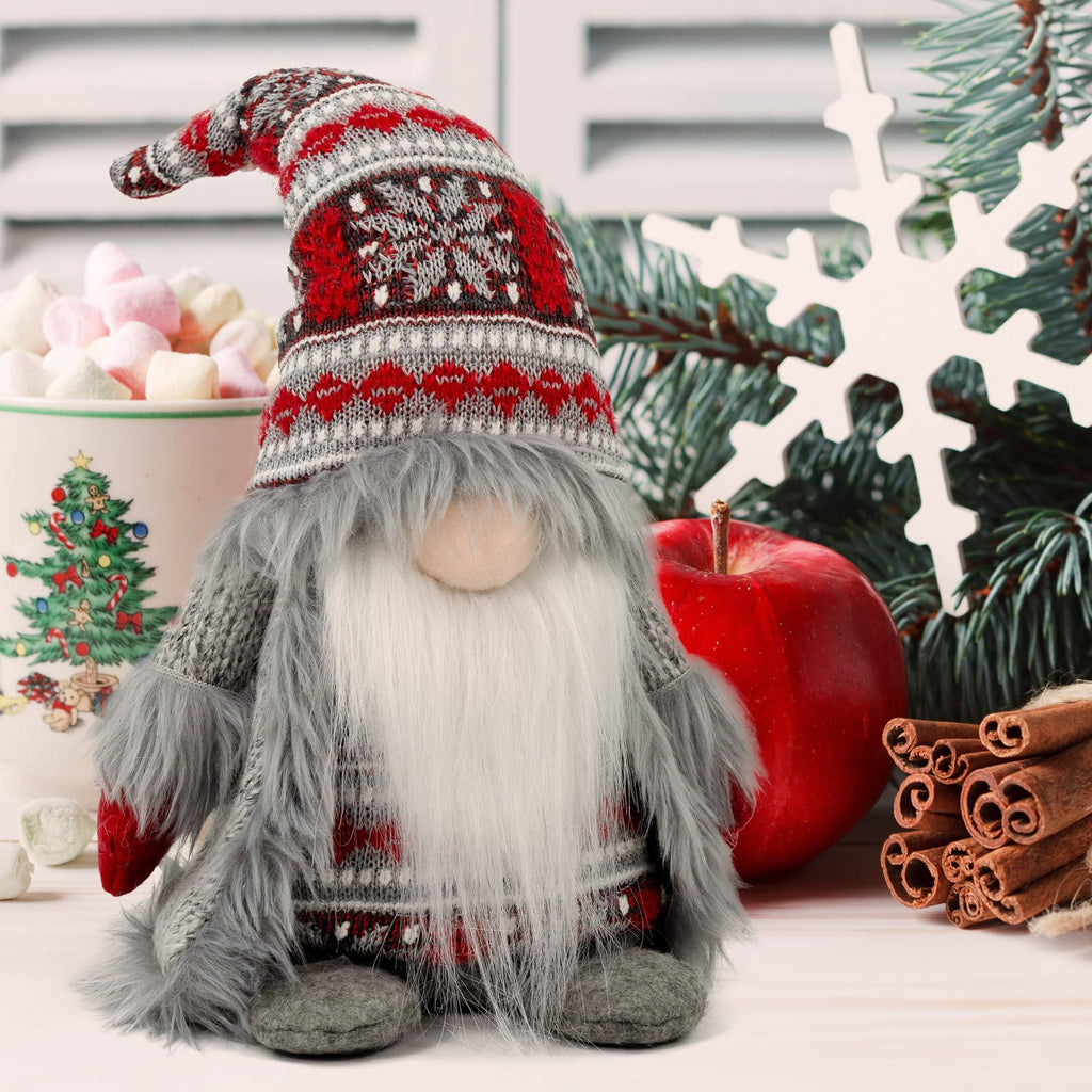 Plush Christmas gnome decoration with gray knit outfit and festive hat, next to a mug of hot chocolate, red apple, cinnamon sticks, and a snowflake ornament.