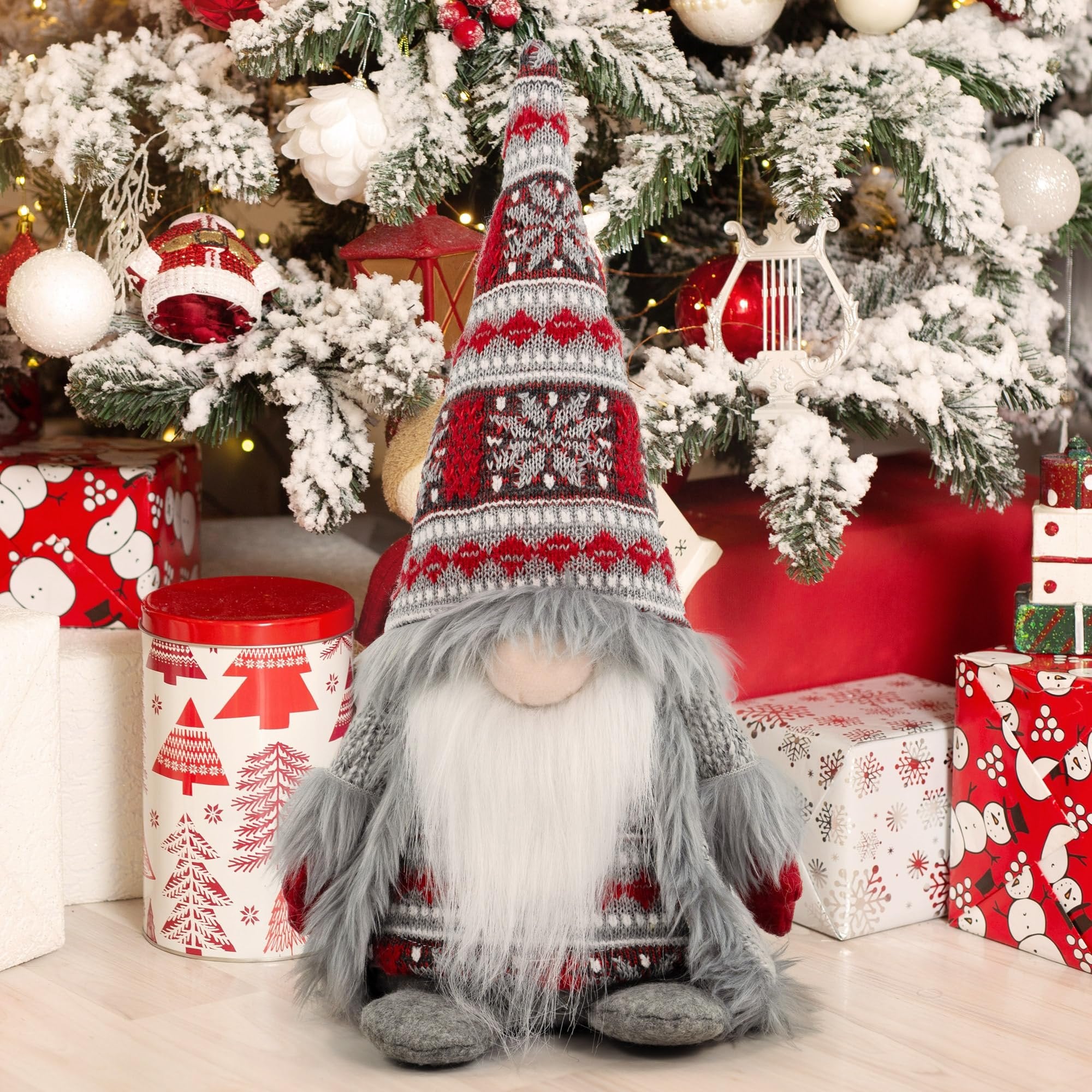 Plush Christmas gnome with gray knit outfit and festive hat sits near a decorated tree with gifts.