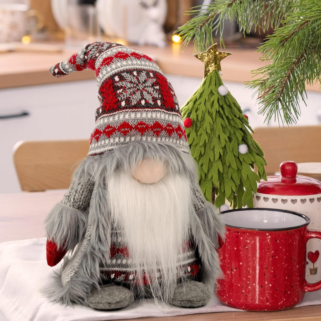 Plush Christmas gnome with gray knit outfit and festive hat sits on a table with a mug and small tree.