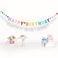 Rainbow 'Happy Birthday' banner with colorful dots, cake, flowers, number 1, and baby booties on display.