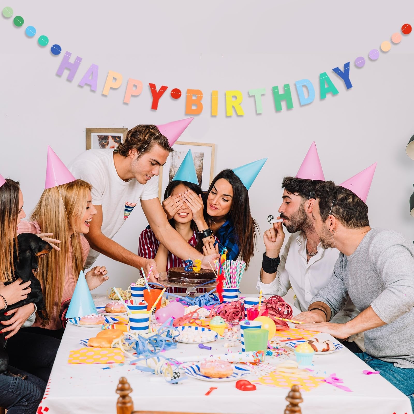 Group of friends celebrating a birthday with a cake and banner, one person covering another's eyes