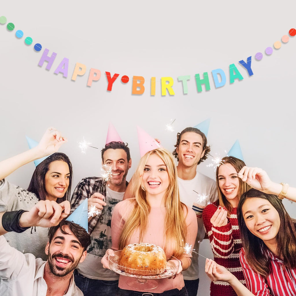 Group of smiling people celebrating a birthday with sparklers and a cake, under a colorful banner