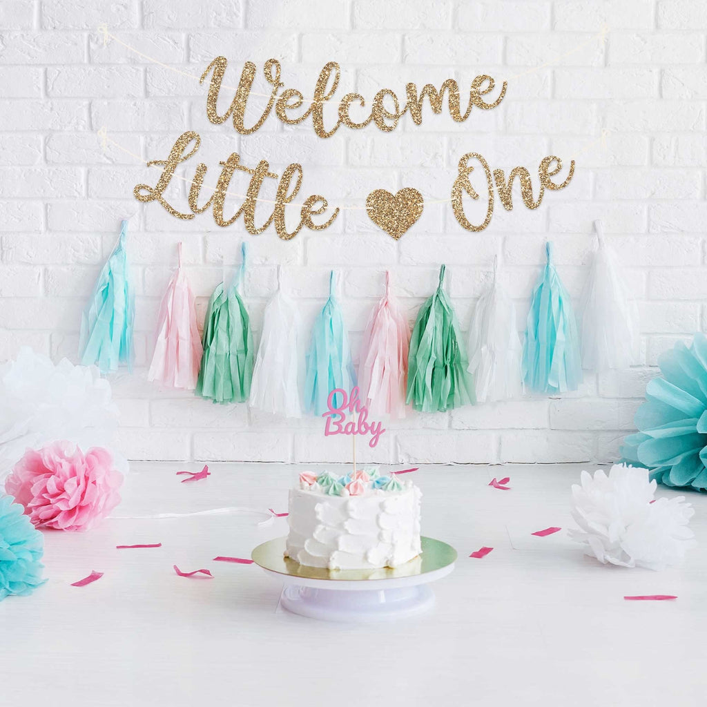 Gold glitter 'Welcome Little One' banner above a cake with 'Oh Baby' topper and pastel tassels.