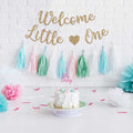 Gold glitter 'Welcome Little One' banner above a cake with 'Oh Baby' topper and pastel tassels.