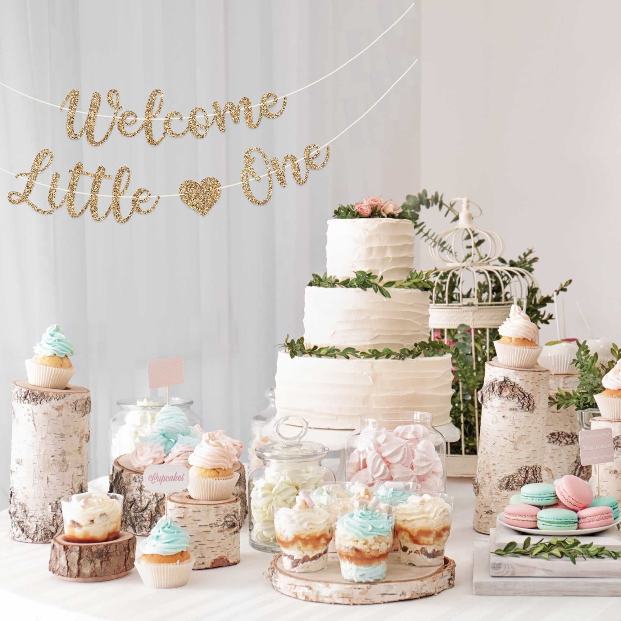 Gold glitter 'Welcome Little One' banner strung across a dessert table with a tiered cake, cupcakes, macarons, and parfaits.