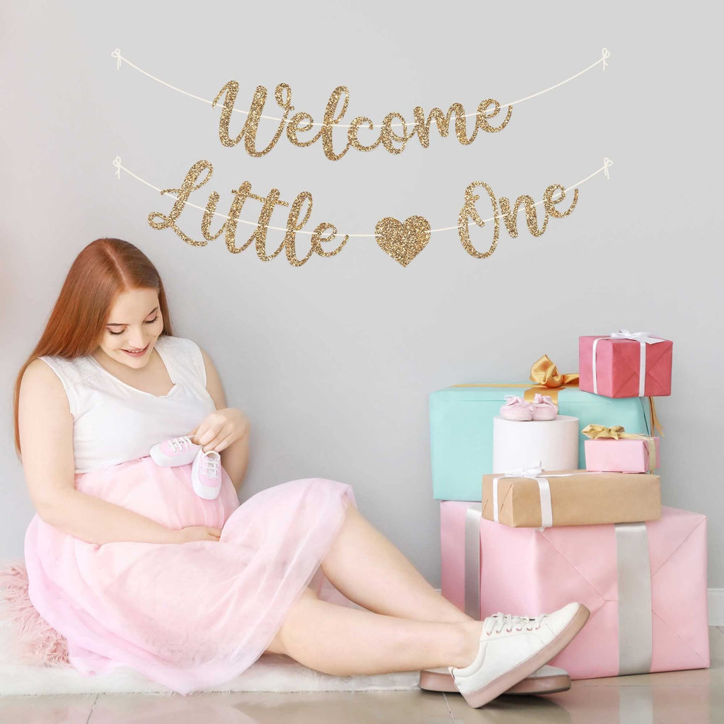 Pregnant woman sitting with baby booties near a 'Welcome Little One' glitter banner and gift boxes