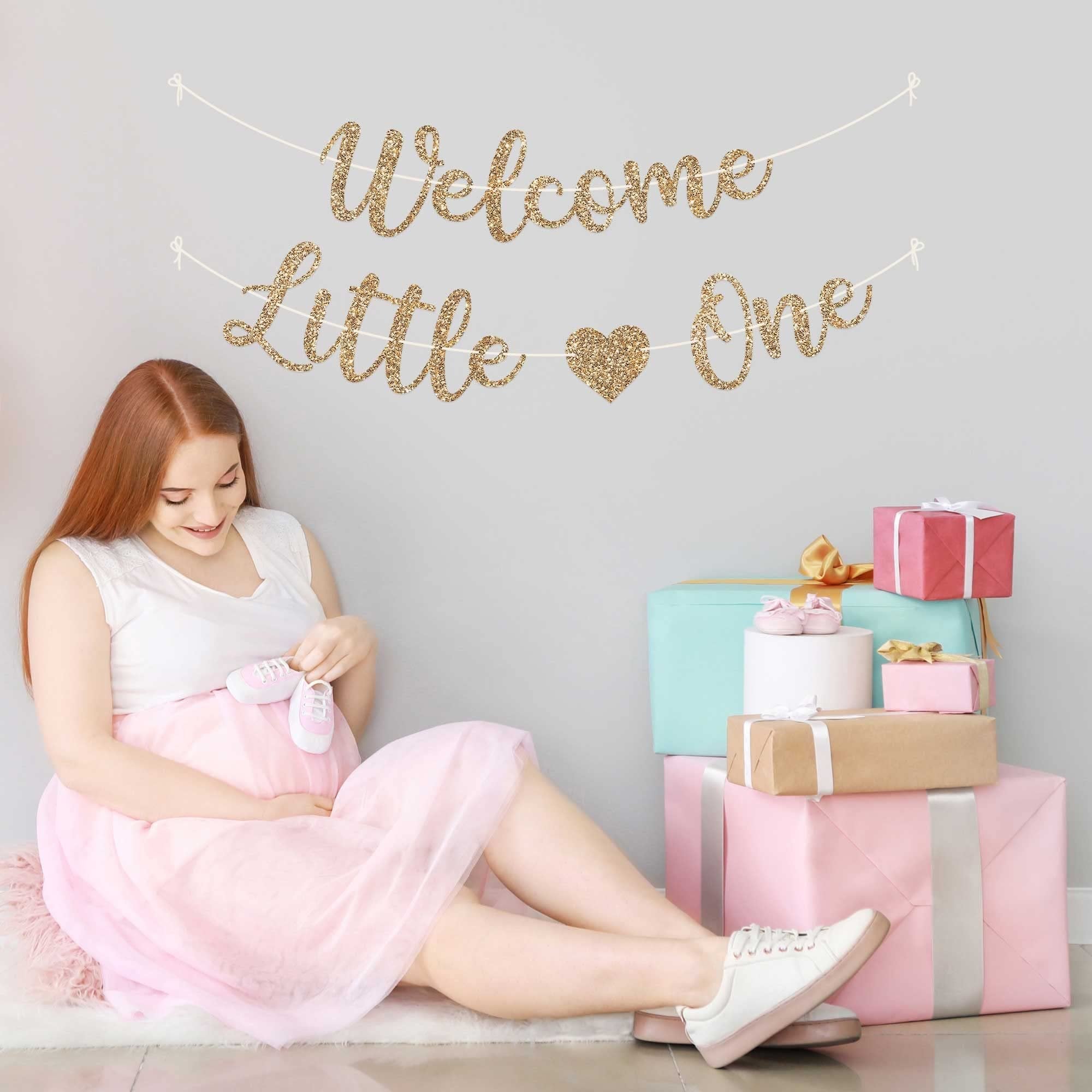 Pregnant woman sitting with baby booties near a 'Welcome Little One' glitter banner and gift boxes