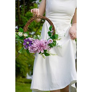 Person in white dress holding a round floral wreath with purple and pink artificial flowers and greenery